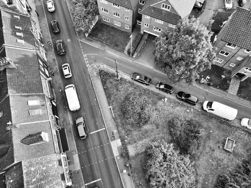 An aerial black-and-white photograph showing a residential street with a row of terraced houses on the left and a small grassy area with trees on the right. Several cars, including a white van, are parked along the curb and driveway in front of the houses. A narrow road runs between the houses and the park, with a few vehicles visible on the street and some parked parallel to the pavement. Near the bottom left, a woman is seen carrying a box, possibly part of a house relocation or packing and moving process managed by Tufnell Park Removals, with a trolley and packing materials such as cardboard boxes and protective blankets nearby. The scene is lit by natural daylight, and the environment appears quiet and organized for a home removal or furniture transport operation.