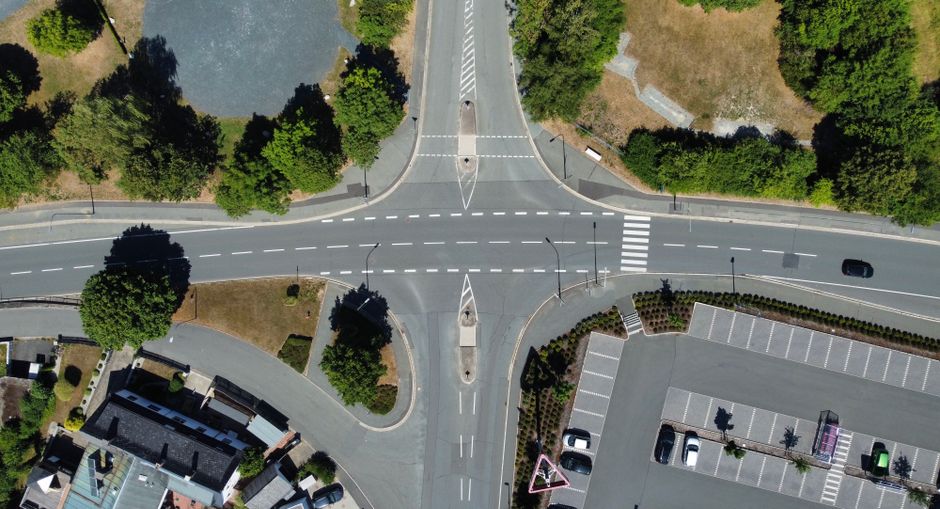 An aerial view of a T-junction intersection in an urban residential area, showing paved roads with white dashed lines marking lanes, pedestrian crosswalks, and traffic islands with greenery and trees. Surrounding the intersection are residential buildings with pitched roofs, a large parking lot with marked parking spaces, and landscaped areas with shrubs and trees. The scene is well-lit, indicating daytime, and no moving vehicles or pedestrians are visible in this moment. The image captures the organized layout of the roads and parking facilities, useful for planning home relocation or furniture transport logistics, which Tufnell Park Removals can assist with as part of their house removals services.